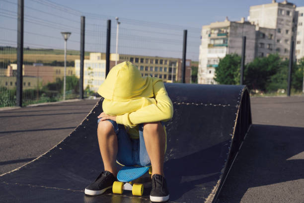 A young boy sits on his skateboard with his eyes covered, portraying a moment of introspection or sadness, contrasting the vibrant outdoor environment around him.