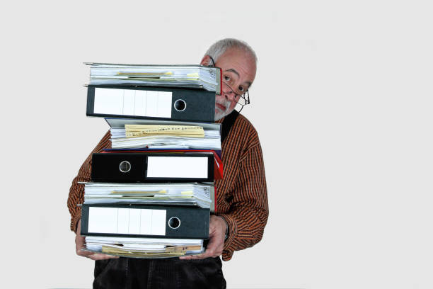 A senior craftsman carries a huge stack of folders, looking pensively to the side. Bureaucracy weighs down businesses and craftsmen, preventing them from focusing on their real work.