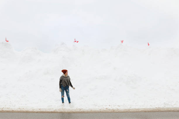 A woman stands in front of a very high snowbank along a lake.