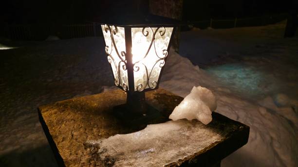 A glowing lantern with ornate metalwork sits on a stone pedestal, surrounded by snow and ice. A small chunk of snow rests nearby, while the night sky and snowy landscape create a serene, cold atmosphere.