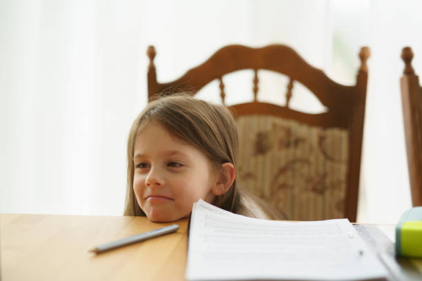 A little tired girl rested her head on the table while doing her homework, sitting in front of a large window behind a vase with flowers. Concept of children's education.