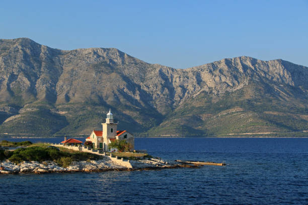 A beautiful summer view of a small stone lighthouse with a red roof, located on a forested peninsula along the Croatian coast. The lighthouse is surrounded by lush pine trees and rocky shoreline, framed by the deep blue waters of the Adriatic Sea and distant mountains under a clear blue sky. This serene landscape captures the peaceful, natural beauty of coastal Croatia and maritime architecture.