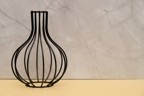 A black vase with a wire frame is placed on a yellow background. The vase is empty and the wire frame is made of metal. The vase has a unique and modern design