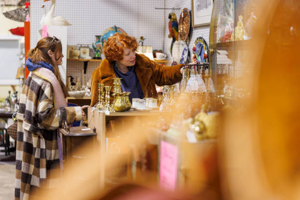 A mature redheaded woman and younger companion carefully examine antique merchandise arranged on pegboard wall displays. They focus on delicate glass bottles, ceramic pieces, and various collectibles spread across multiple shelving levels in this well-organized vintage goods store