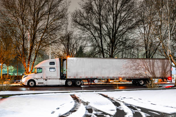istock Professional white big rig semi truck with turned on headlight and refrigerated semi trailer standing on the winter city street with snow and ice at night time 2201405492