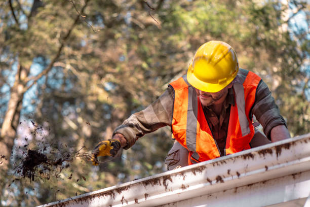 Worker in safety gear and yellow hard hat performs roof maintenance. Contractor cleans gutters on residential house