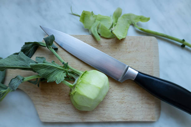 Peeled fresh kohlrabi rests on a cutting board, with its removed peel placed nearby. A knife is positioned beside the vegetable, ready for further preparation.