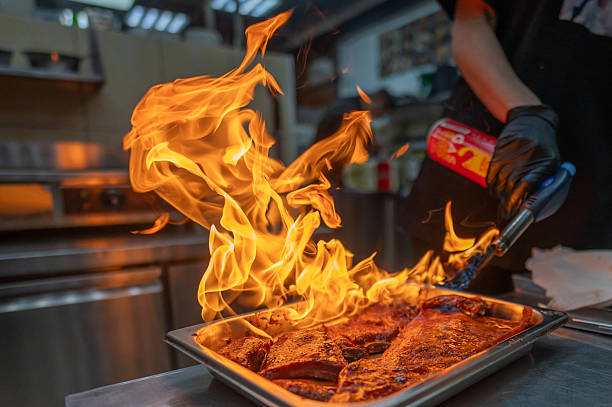 A chef skillfully uses a culinary torch to sear juicy meat, creating dramatic flames in a vibrant kitchen setting. The intense heat highlights the delicious texture and spices
