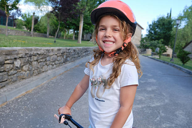 Portrait of cheerful little girl in casual clothes and helmet smiling and looking at camera while riding push scooter on street during daytime