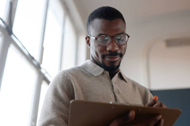 istock Adult Black Man with Glasses Writing on Clipboard while Working in Office 2201460168