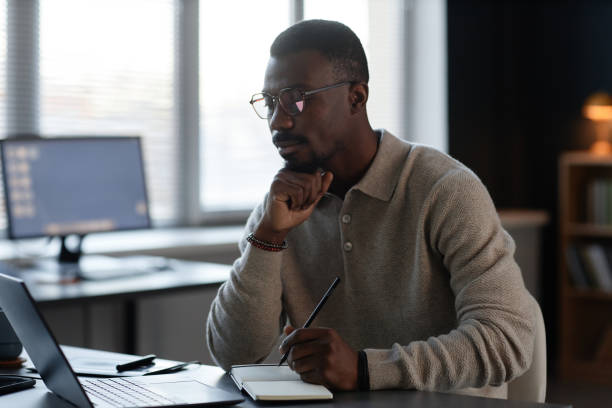 istock Focused Black Man Writing in Notebook while Working on Laptop at Desk in Office 2201460175