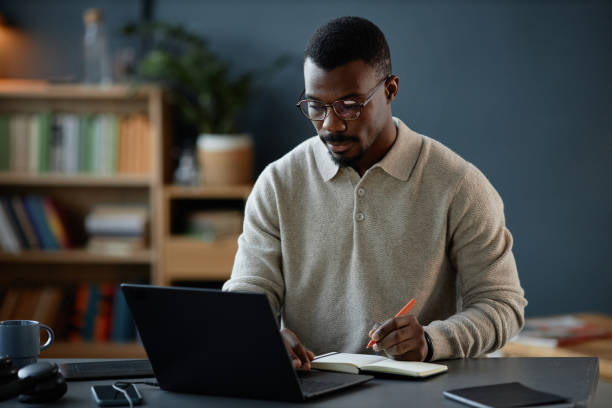 istock Adult Black Man Writing in Notebook while Using Laptop at Desk in Office 2201460184