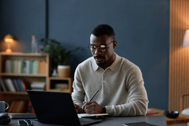 istock Black Male Interior Designer Writing in Notebook while Using Laptop at Desk in Office 2201460185