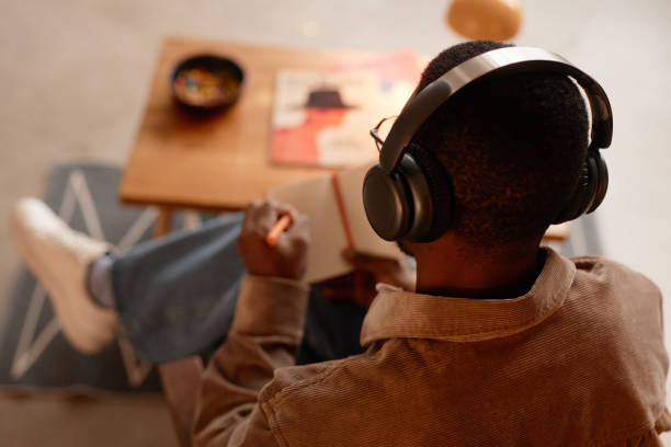 Black Man Listening to Podcast Wearing Headphones while Writing Notes in Home Office