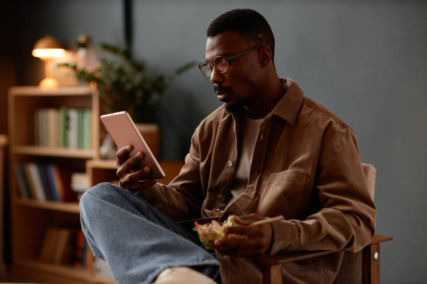 istock Adult Black Man Watching Video on Tablet during Lunch Break in Home Office 2201460238