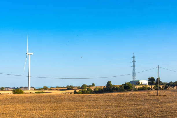A wind turbine and electrical power lines stand in a dry rural landscape under a clear blue sky. The scene highlights the integration of renewable energy and traditional infrastructure.