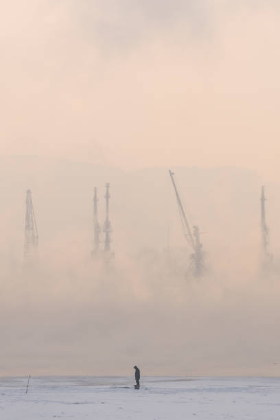 A lone fisherman stands in a misty industrial scene that evokes tranquility and reflection. A solitary figure stands on the icy waters, surrounded by industrial cranes shrouded in thick fog and mist.