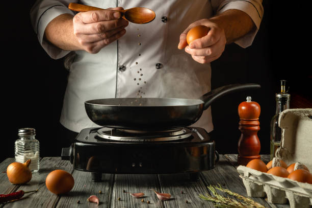istock A skilled chef expertly holds an egg above a hot frying pan and spoon, surrounded by ingredients like spices and herbs on a rustic wooden table. The atmosphere hints at culinary creativity and passion 2201482853