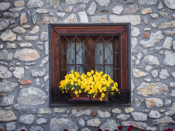 A charming wooden window with wrought iron bars set in a rustic stone wall, adorned with vibrant yellow flowers in a planter. The textured stone and warm wood create a cozy, traditional ambiance.