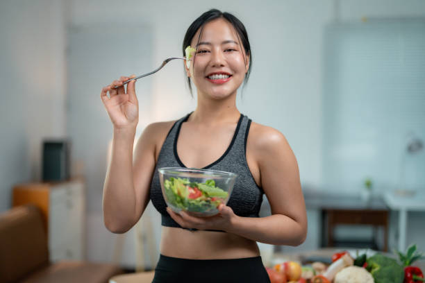 istock Young asian sportswoman eating fresh green salad, smiling while holding fork, wearing athletic clothing in home kitchen setting 2201499134