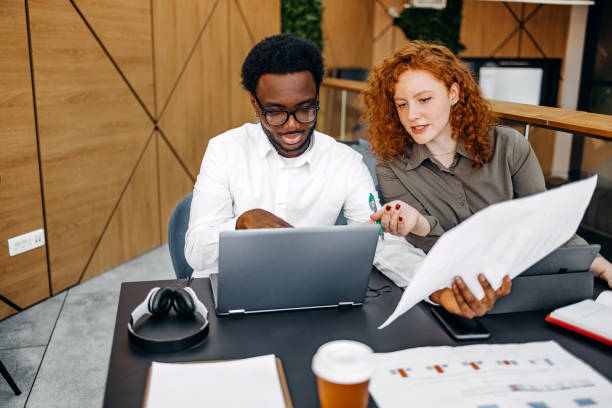 istock Two young adults working together in office while sitting in front of laptop and digital tablet and looking at charts 2201501067