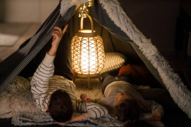 A young brother and sister enjoy a quiet and intimate moment inside a homemade blanket fort in their bedroom. The boy, dressed in a striped sweater, sits cross-legged, engaging in a heartfelt conversation with his younger sister, who listens attentively.