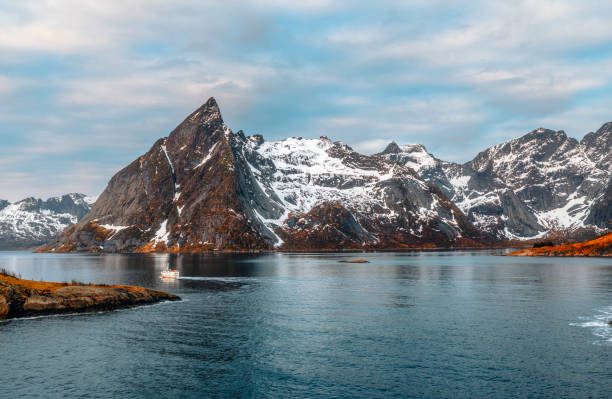 The majestic Olstinden Mountain dominates the Arctic landscape, towering over the calm waters of the fjord in Hamnoy, Norway. The golden sunlight casts a warm glow on the rugged, snow-dusted slopes, creating a striking contrast against the icy blue sky. This iconic peak, a defining feature of the Lofoten Islands, is reflected in the serene waters below, enhancing the dramatic scenery of Norway’s remote wilderness