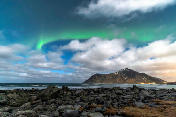 The mesmerizing Northern Lights dance across the night sky over Skagsanden Beach in Lofoten, Norway. The vibrant green aurora illuminates the snow-covered mountain range near Ramberg, reflecting off the coastal waters. Wispy clouds add depth to the Arctic night, while the rugged shoreline and scattered rocks enhance the dramatic scenery. A breathtaking winter landscape showcasing the magic of the aurora borealis in the far north
