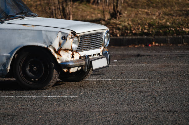 A weathered, abandoned car with rust, dents, and a severely damaged front end sitting in an empty parking lot. The worn-out tires and peeling paint highlight the passage of time and neglect.