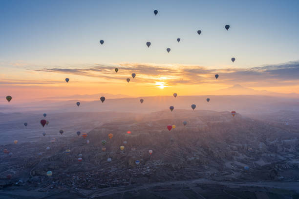 Aerial view of numerous hot air balloons flying over rock formations and town in Cappadocia, Turkey, against morning sun