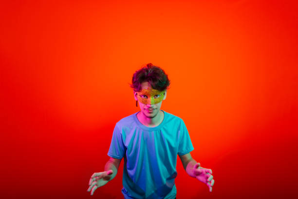 istock Happy handsome young man wearing carnival mask looking and making hand gestures to the camera. Isolated on orange neon background. 2201542334