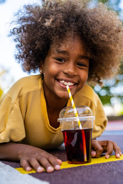 A cheerful African American preschool girl with a natural afro lies on her stomach, while smiling brightly. In front of her sits a plastic cup filled with grape juice, complete with a straw, adding to the playful and carefree moment.