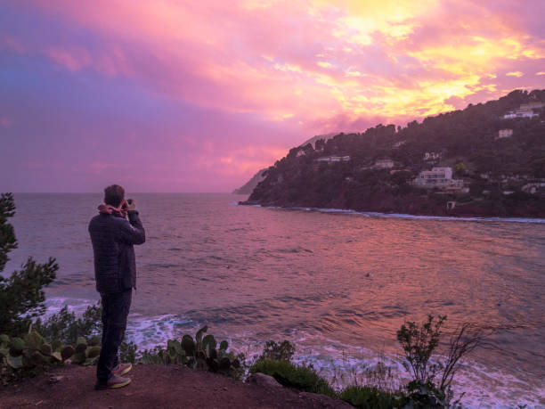 Photographer takes photos on edge of sea at sunset across from village, dramatic light in sky