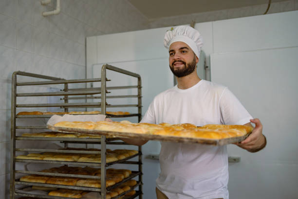 Young male baker smiling and holding a tray of freshly baked bread, standing in front of a metal rack filled with more bread in a bakery kitchen