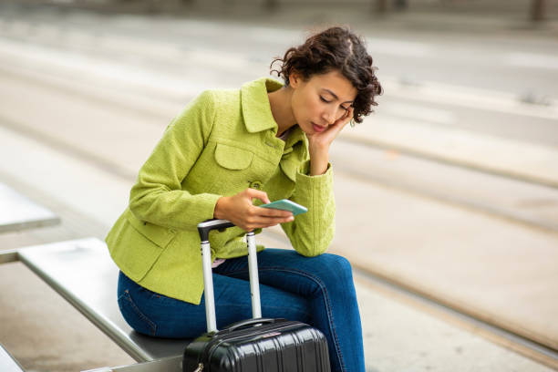 istock Sad woman using mobile phone while waiting with luggage at bus stop 2201572996