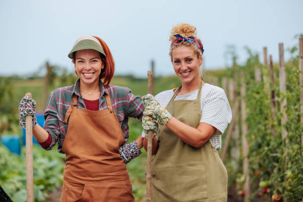 Two young adult women happily working in a vegetable garden, holding tools and wearing aprons, surrounded by lush greenery and healthy plants