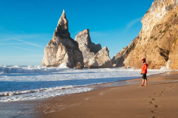 istock Lonely woman dressed red jacket walking by Beautiful abandoned Atlantic ocean Ursa beach and enjoying coastal surf with foamy waves crashing on rocky cliffs on Winter February coast in Portugal. 2201594002