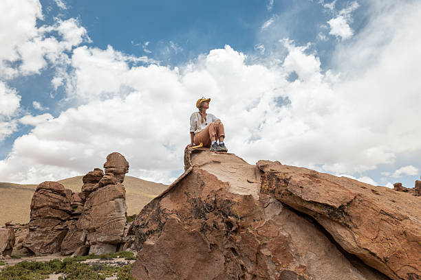 A traveler in casual attire sits atop a large rock, admiring the vast desert landscape with unique rock formations and a cloudy blue sky in the background.