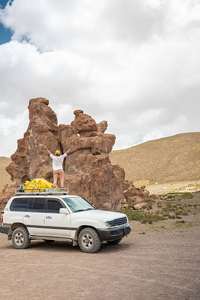 A woman in casual attire stands near towering rock formations, next to an off-road vehicle loaded with gear, enjoying an adventurous journey through a remote desert.