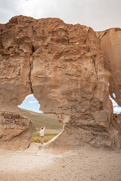 A traveler stands beneath a towering eroded rock formation with natural openings, admiring the unique geological structure in a remote desert landscape.