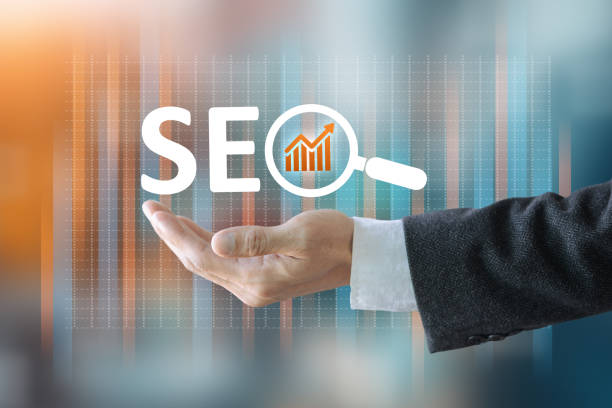 A person is holding a magnifying glass over the word SEO, which stands for Search Engine Optimization. The person is likely trying to emphasize the importance of SEO in digital marketing