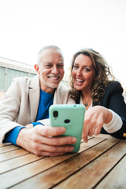 istock Vertical. Middle aged couple having fun sharing media with a cellphone. Mature people watching the smartphone on a coffee shop or bar. Mid adult friends buying on a market place using a mobile phone 2201604404
