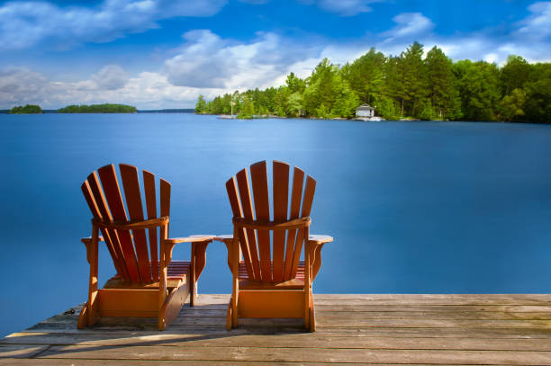 istock Adirondack chairs on a dock facing a calm lake and white cottage. 2201609348