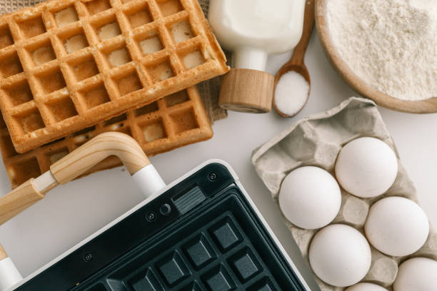 istock Ingredients for homemade waffles arranged on a white surface, including flour, sugar, eggs, and milk. A waffle maker with wooden handles is visible in the frame 2201645112