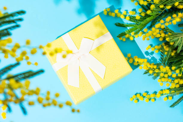 istock Yellow gift box on blue background with mimosa flowers and sunlight. 2201664635