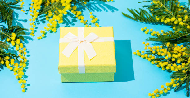 istock Yellow gift box on blue background with mimosa flowers and sunlight. 2201664641