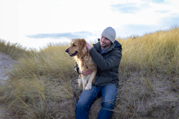 A wide shot of a man sitting down with his golden retriever dog while on a walk at Druridge Bay in Northumberland. The man is looking at his dog while smiling, and the dog sitting on the sand bank with his paws on his legs. He is wearing casual, warm clothing, as it is winter.\n\nVideos are available similar to this scenario.