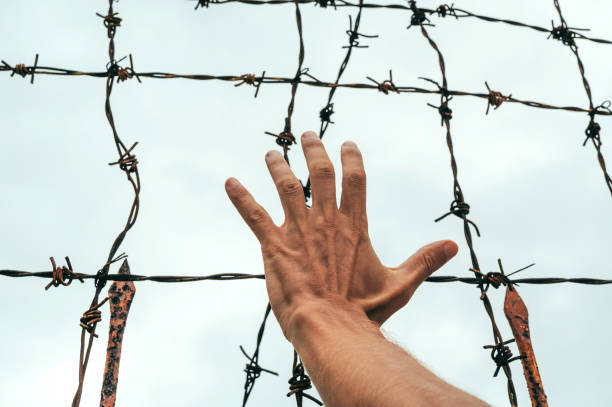 istock men's hand behind a barbed wire fence against the sky. 2201668297