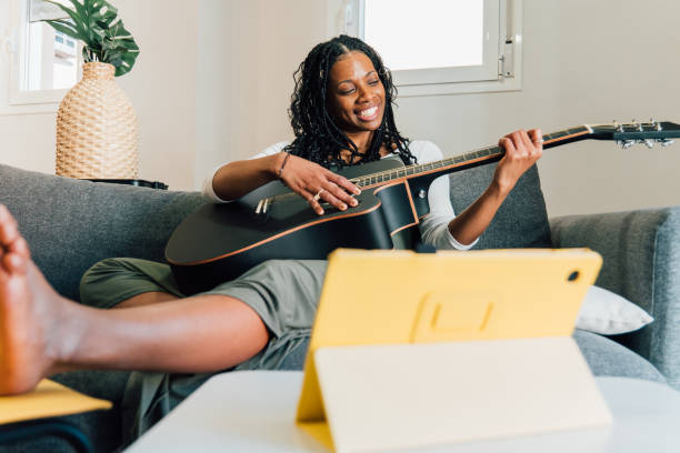 istock Black woman sitting on sofa, strumming acoustic guitar while glancing at digital tablet 2201670790