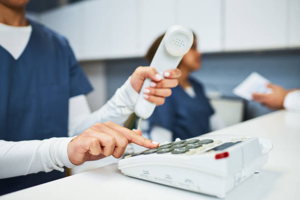 Healthcare workers in uniforms interact within a medical facility. Focus is on a receptionist dialling on a landline phone, conveying professionalism and effective communication in a healthcare environment.